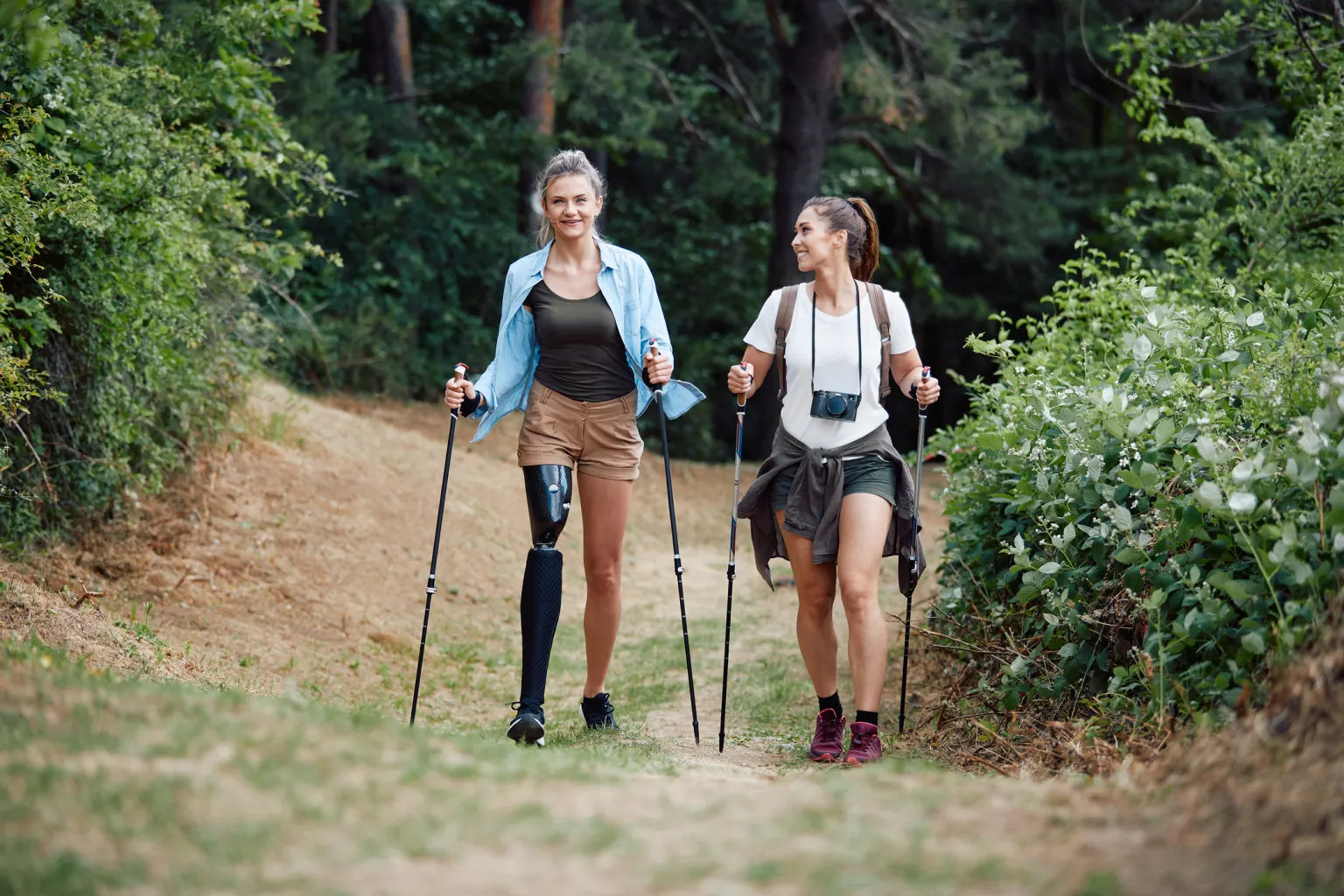 Zwei Frauen gehen mit Wanderstöcken einen Waldweg entlang und unterhalten sich. Die linke trägt eine Beinprothese.