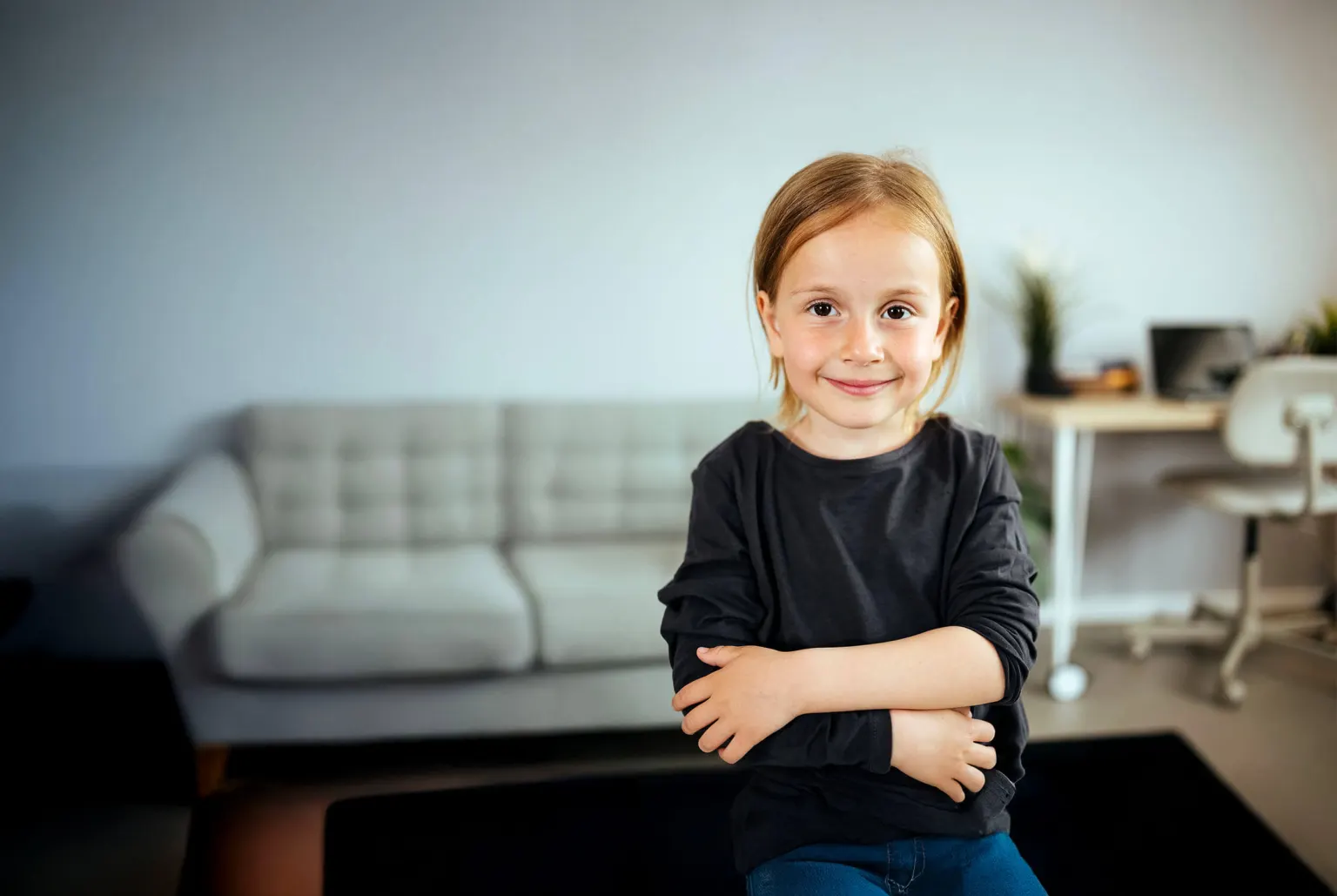 Ein Kind sitzt mit verschränkten Armen im Wohnzimmer und lächelt in die Kamera.