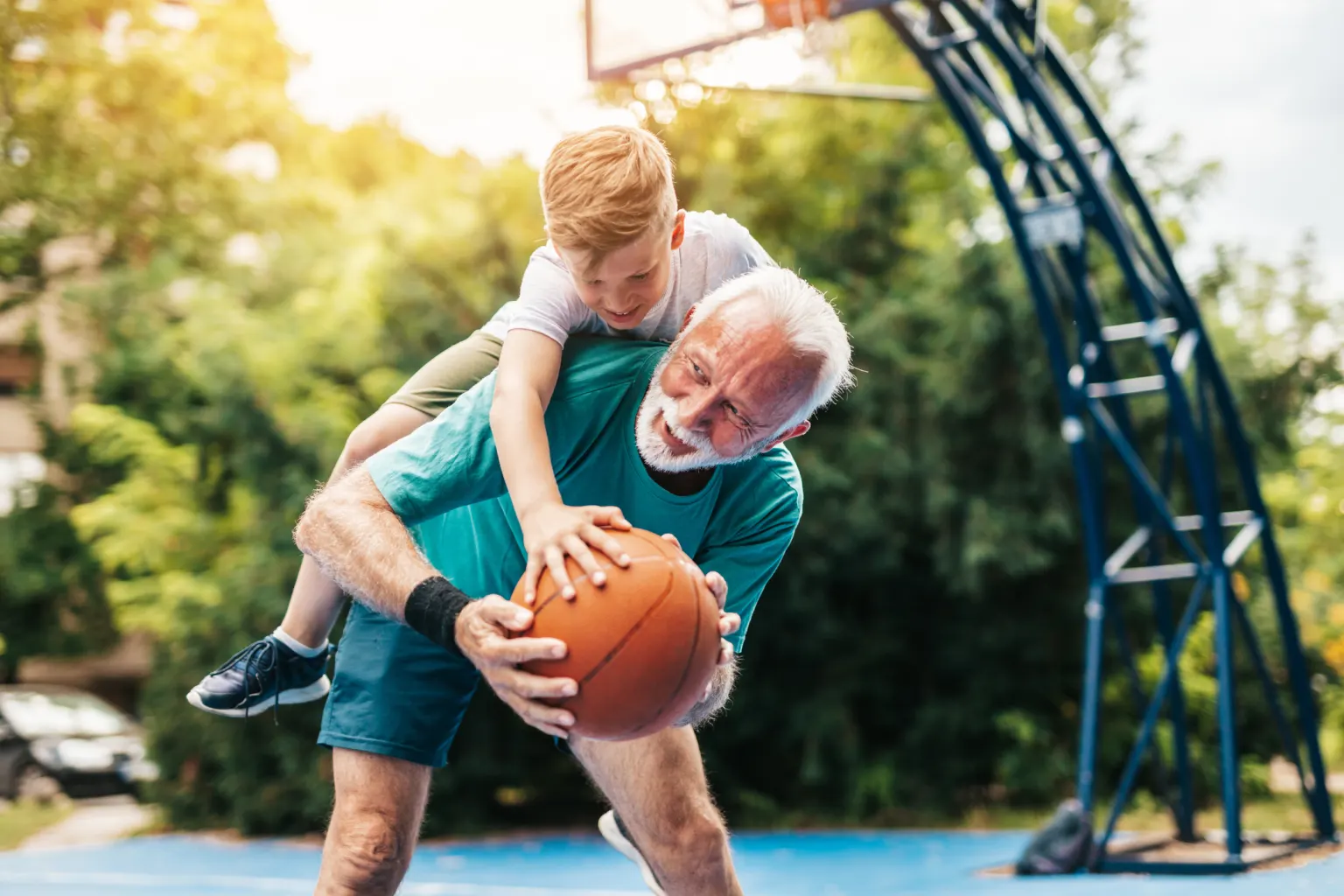 Ein älterer Mann spielt auf einem Basketballplatz mit seinem Enkel. Der Junge liegt lachend über seiner Schulter, der Mann hält den Ball.
